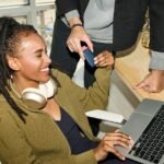 Smiling woman uses laptop, person holds credit card nearby