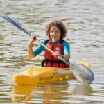 Enfant rieur dans kayak jaune, pagaie à l'eau, gilet orange