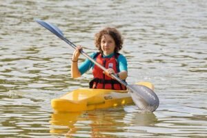 Enfant rieur dans kayak jaune, pagaie à l'eau, gilet orange