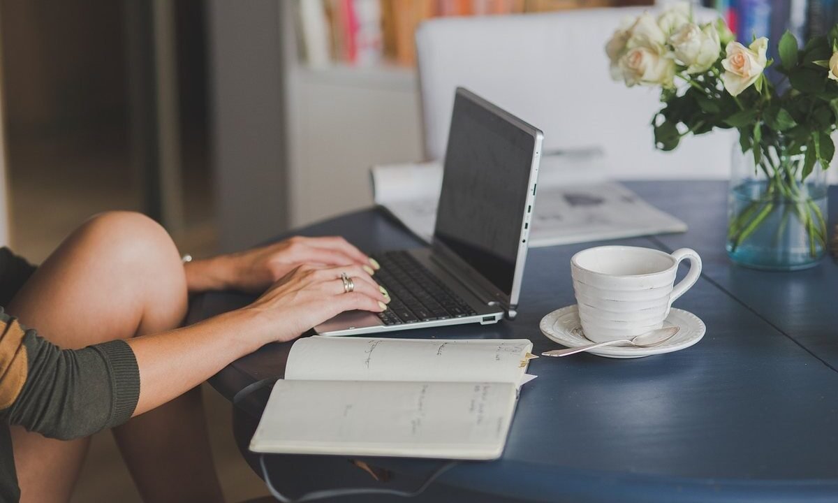 Une main tapant sur un ordinateur portable, cahier ouvert, tasse et fleurs sur table