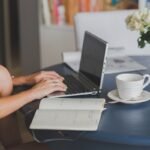 Une main tapant sur un ordinateur portable, cahier ouvert, tasse et fleurs sur table