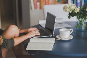 Une main tapant sur un ordinateur portable, cahier ouvert, tasse et fleurs sur table