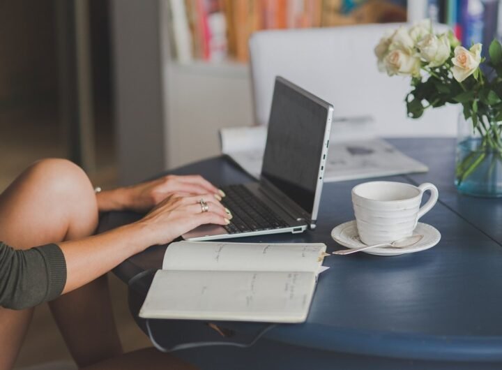 Une main tapant sur un ordinateur portable, cahier ouvert, tasse et fleurs sur table